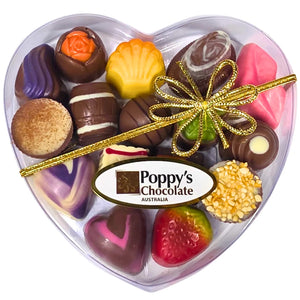 A heart-shaped clear gift box containing an assortment of gourmet chocolates, tied with a Gold ribbon and branded with 'Poppy's Chocolate'.