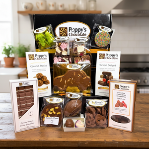 A Poppy's Chocolate gift assortment displayed on a wooden table in a kitchen setting, featuring multiple packaged chocolate items, a large black gift box, and printed flavor cards including Coconut Crisp, Turkish Delight, and Chocolate Covered Pistachio.