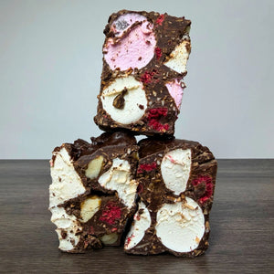 Three stacked square pieces of chocolate-covered nut and marshmallow bark on a dark wooden surface against a light gray background.