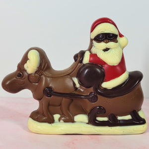 Chocolate Santa wearing sunglasses and a red suit, riding in a sleigh behind a reindeer, with white chocolate details.