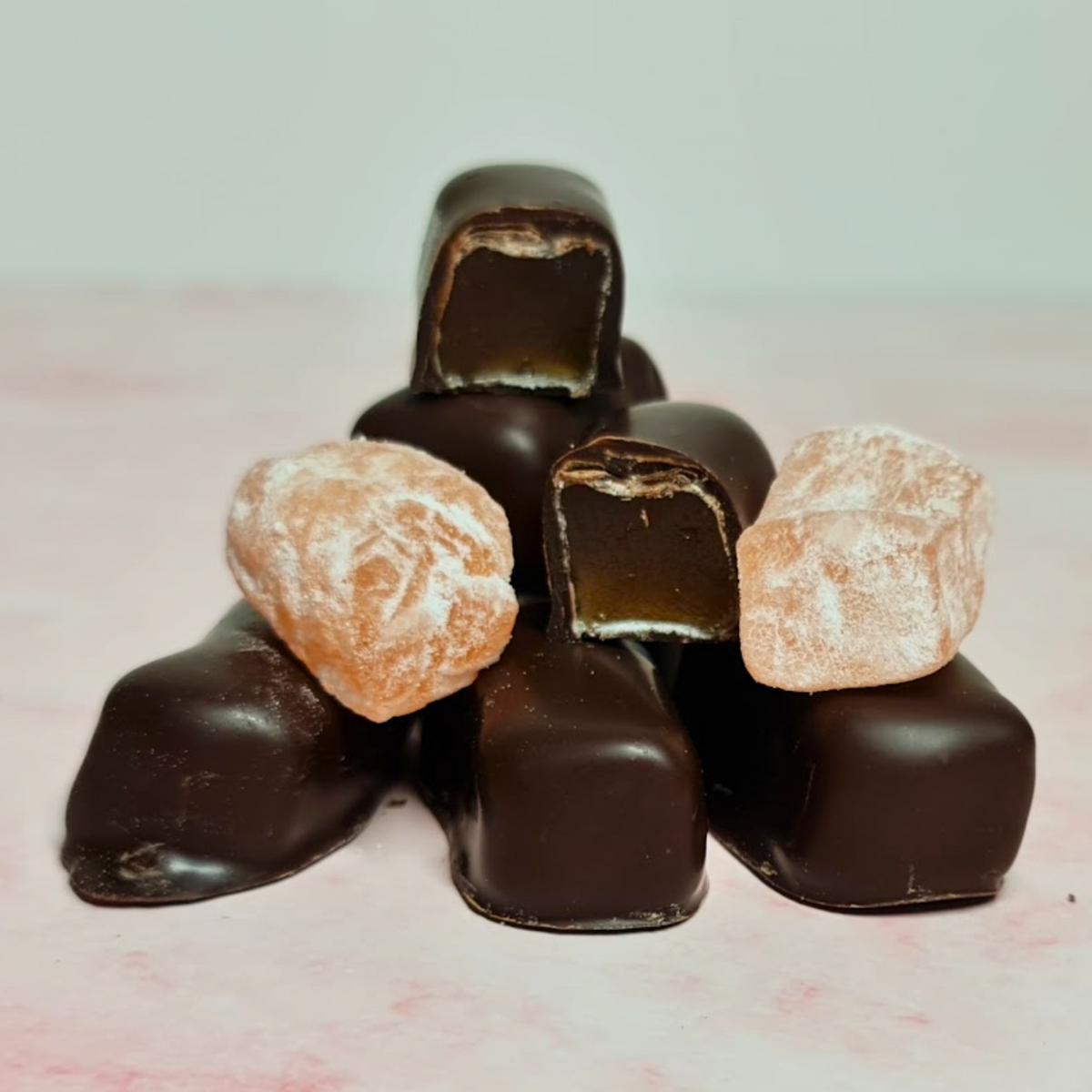 A tempting pyramid of Poppy&#39;s Chocolate Coated Turkish Delight Dark Chocolate 100g chocolates, with one piece split in half to showcase its caramel-like center, sits attractively on the soft pink surface—an ideal gift. Nearby lie two powdered sugar-coated candies evoking the traditional taste of Turkish Delight.