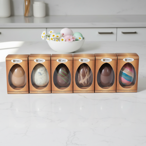 Six easter eggs in boxes with windowed lids on a white countertop, with a bowl of Easter eggs in the background.