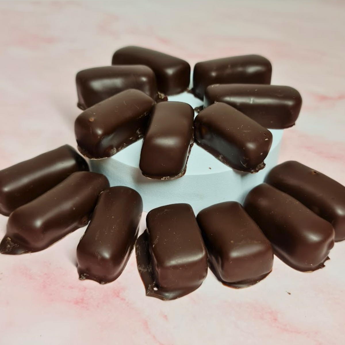 A circle of Poppy&#39;s Chocolate Coated Turkish Delight Dark Chocolate 100g is arranged neatly around a small blue platform on a pink marbled surface, making it perfect for gifting.