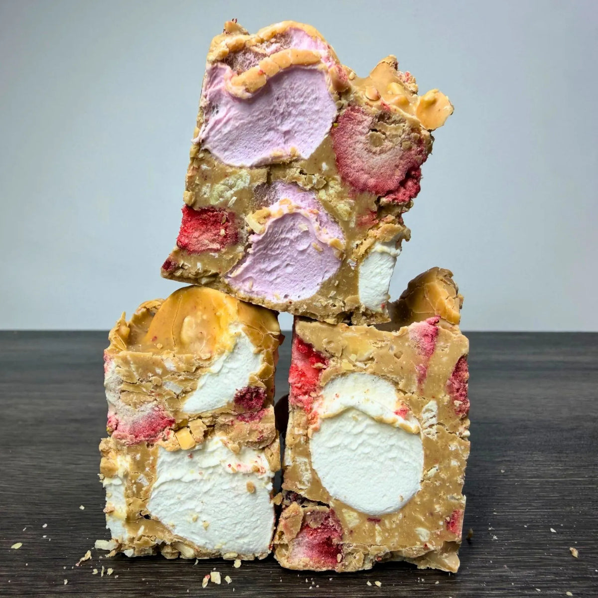 Three stacked pieces of chocolate malt bark with visible swirls of lavender ice cream, white ice cream, pink strawberry pieces, and caramel-like coating on a dark wooden surface against a light gray background.