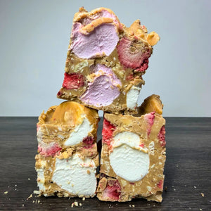 Three stacked pieces of chocolate malt bark with visible swirls of lavender ice cream, white ice cream, pink strawberry pieces, and caramel-like coating on a dark wooden surface against a light gray background.