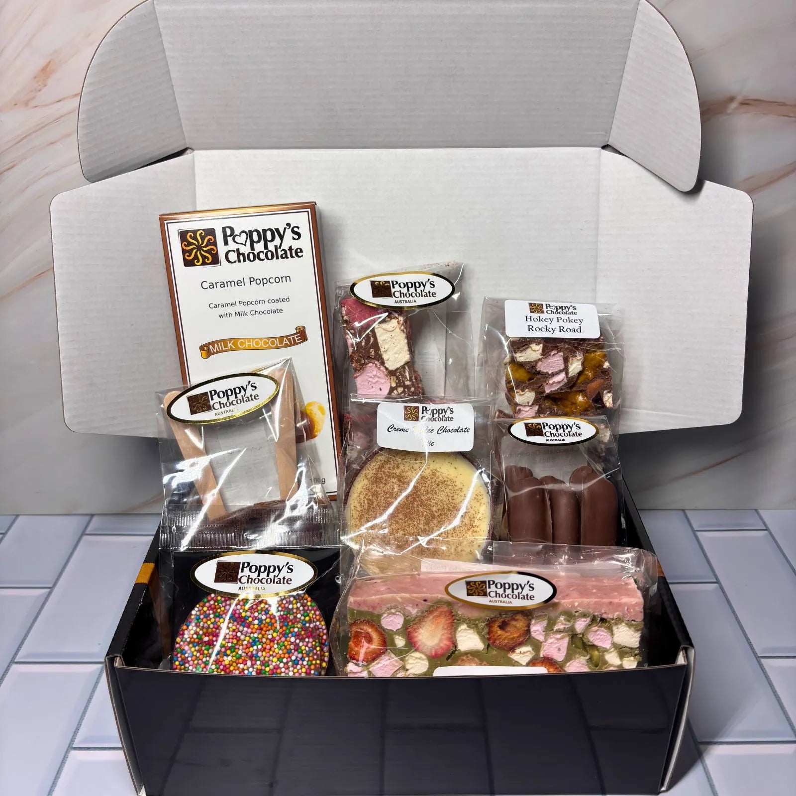 A chocolate gift basket assortment from Poppy's Chocolate featuring various packaged treats including Rocky Road chocolates, milk chocolate, caramel popcorn, and a large black gift box on a white background.